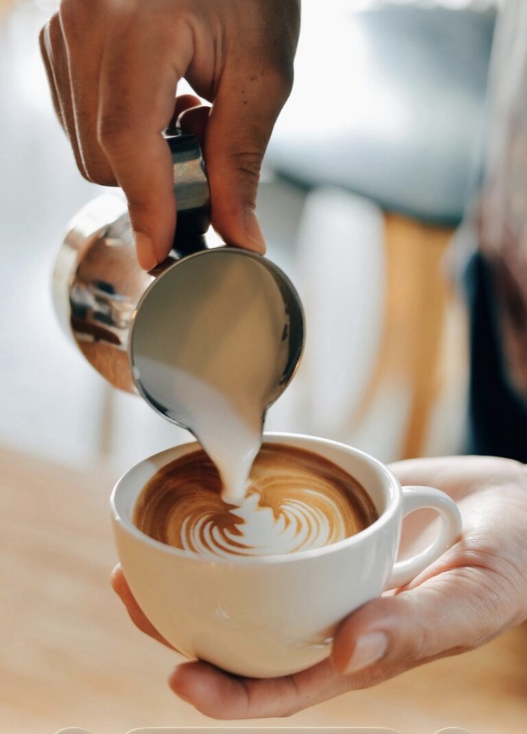 A hand pours steamed milk into a coffee cup, creating a swirling latte art design on the surface of the rich espresso.