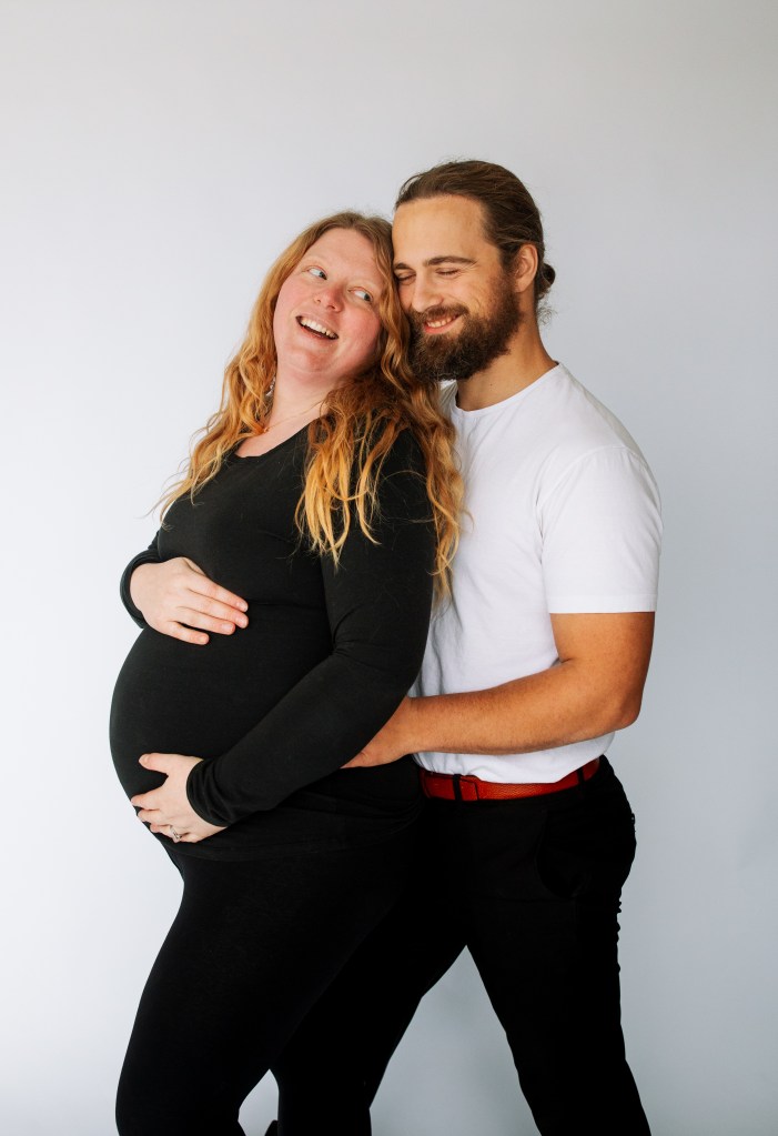 A smiling pregnant woman cradles her belly, embraced from behind by a man. They share a joyful moment against a simple gray background.