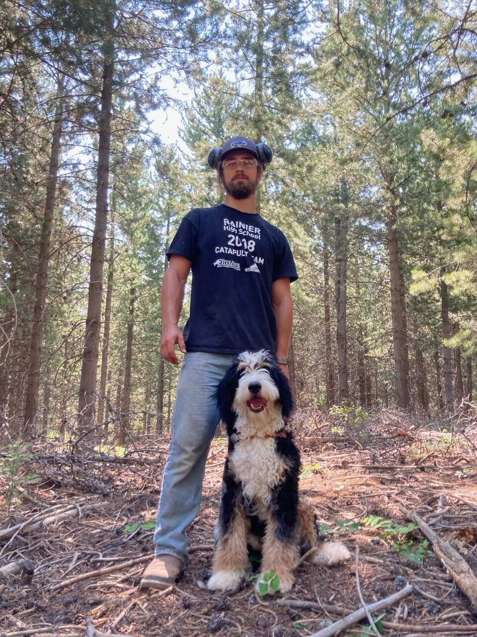 Man stands in a forest, wearing a t-shirt and headphones, next to a smiling black and white dog. The scene highlights their outdoor companionship.